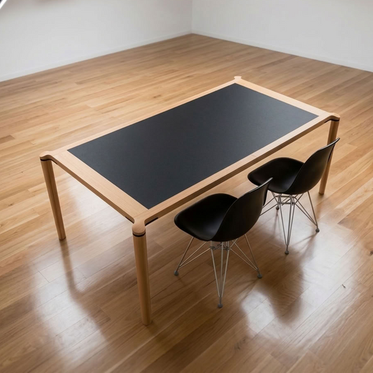A modern rectangular dining table with a light natural wood frame and a black rectangular center panel, shown with two black chairs on a polished wood floor in a studio-like room.